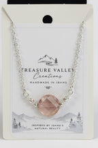 A silver chain necklace with a faceted blush pink gemstone pendant, displayed on its white backing card. The card features the brand name 'Treasure Valley Creations' and a scenic Idaho graphic.