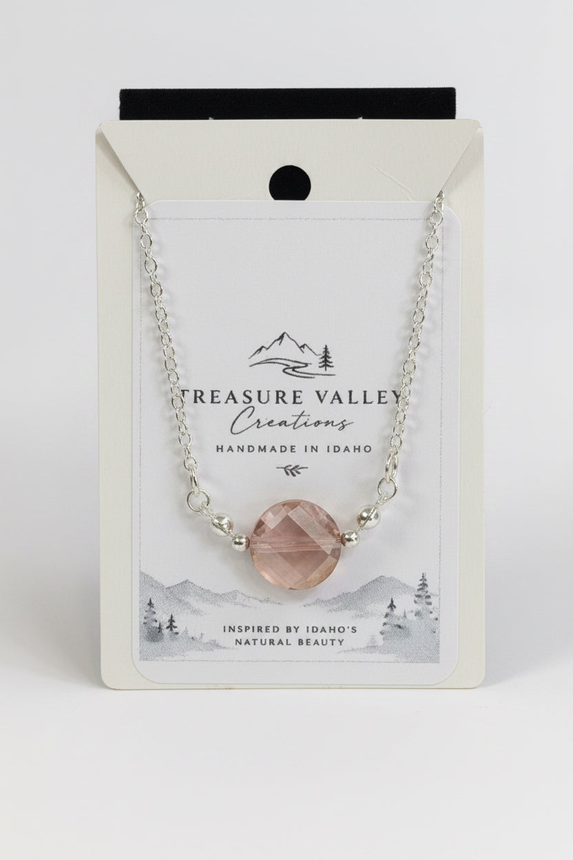 A silver chain necklace with a faceted blush pink gemstone pendant, displayed on its white backing card. The card features the brand name 'Treasure Valley Creations' and a scenic Idaho graphic.