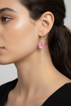 A close-up side profile of a woman wearing a dangling earring with a pink square stone and a silver chain.