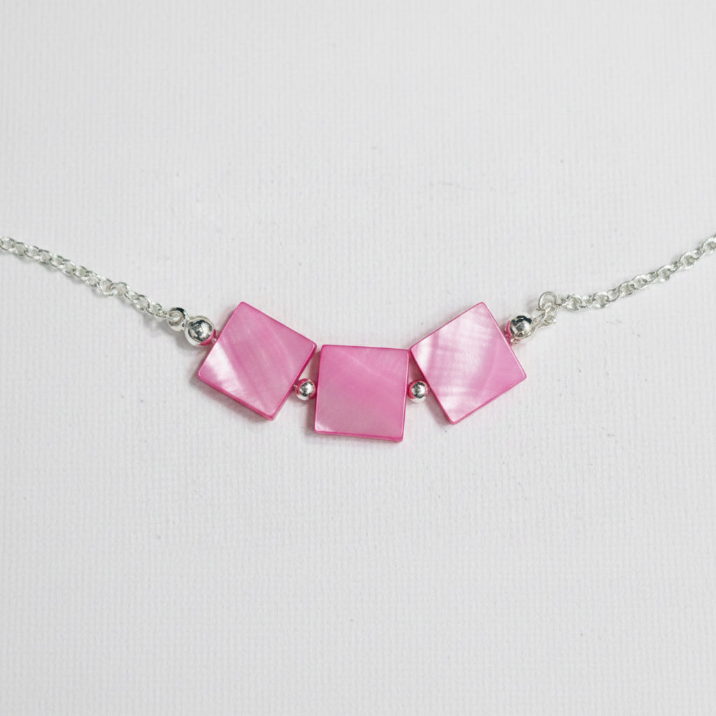 A close-up of a necklace featuring three square-shaped pink mother-of-pearl beads on a silver chain, laid flat on a light gray textured surface.