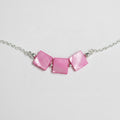 A close-up of a necklace featuring three square-shaped pink mother-of-pearl beads on a silver chain, laid flat on a light gray textured surface.