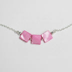 A close-up of a necklace featuring three square-shaped pink mother-of-pearl beads on a silver chain, laid flat on a light gray textured surface.