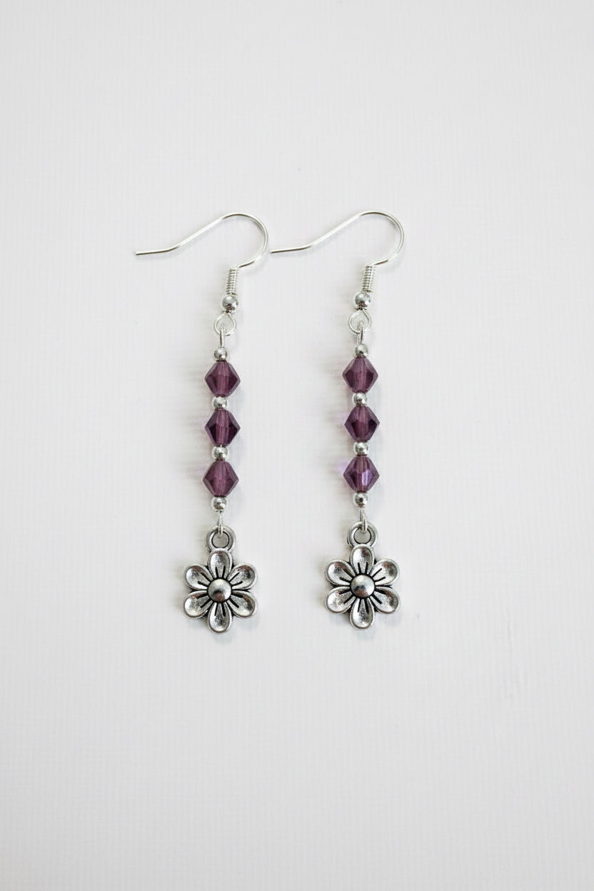 A pair of silver dangle earrings featuring three faceted purple beads and a small silver flower charm at the bottom, shown against a plain light background.