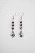 A pair of silver dangle earrings featuring three faceted purple beads and a small silver flower charm at the bottom, shown against a plain light background.