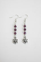 A pair of silver dangle earrings featuring three faceted purple beads and a small silver flower charm at the bottom, shown against a plain light background.