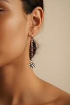 A close-up of a model's ear wearing a long silver dangle earring with small dark purple beads and a dangling flower charm, against a warm beige background.