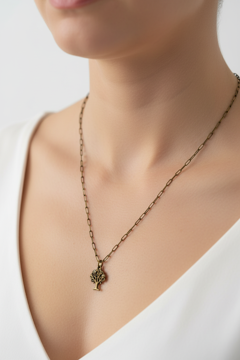 A close-up of a model wearing a delicate antique bronze chain necklace with a small tree of life pendant, styled with a white V-neck top against a soft, light background.
