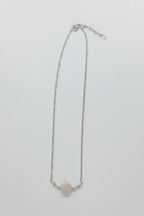 A delicate silver chain necklace featuring a small, pale pink rose quartz crystal pendant, displayed against a neutral light gray background.