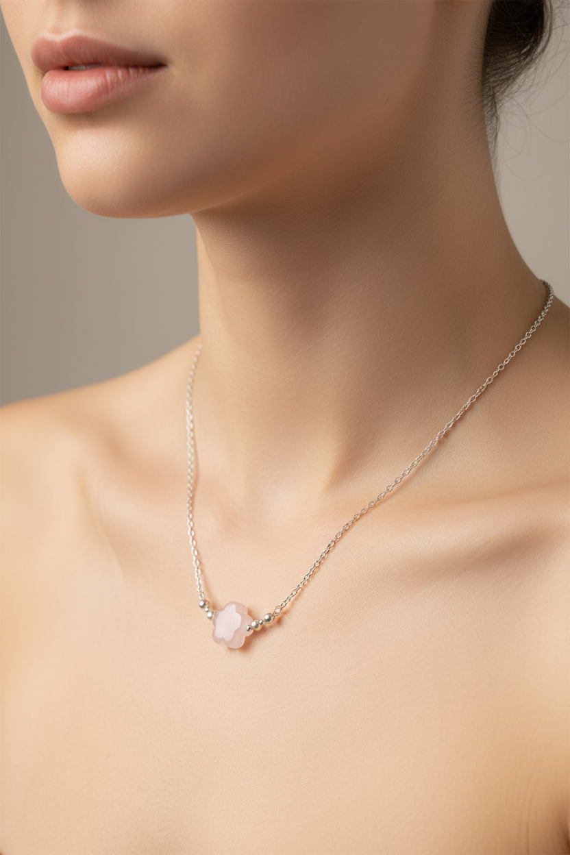 A close-up of a model's neck and upper chest wearing a delicate necklace with a pale pink flower-shaped pendant on a fine chain.