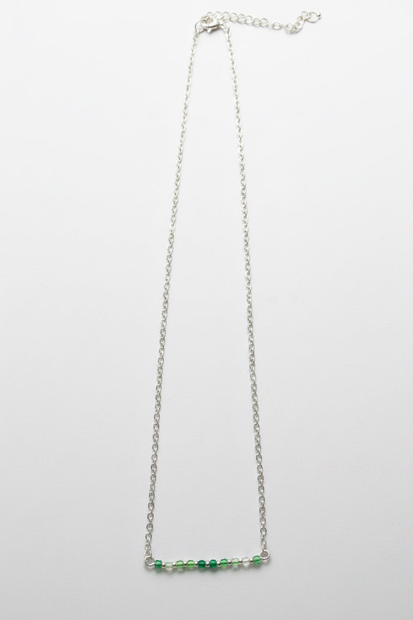 A delicate silver chain necklace featuring a short horizontal bar at the bottom adorned with small green and clear faceted beads, shown against a plain light gray background.