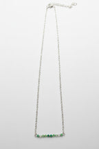 A delicate silver chain necklace featuring a short horizontal bar at the bottom adorned with small green and clear faceted beads, shown against a plain light gray background.