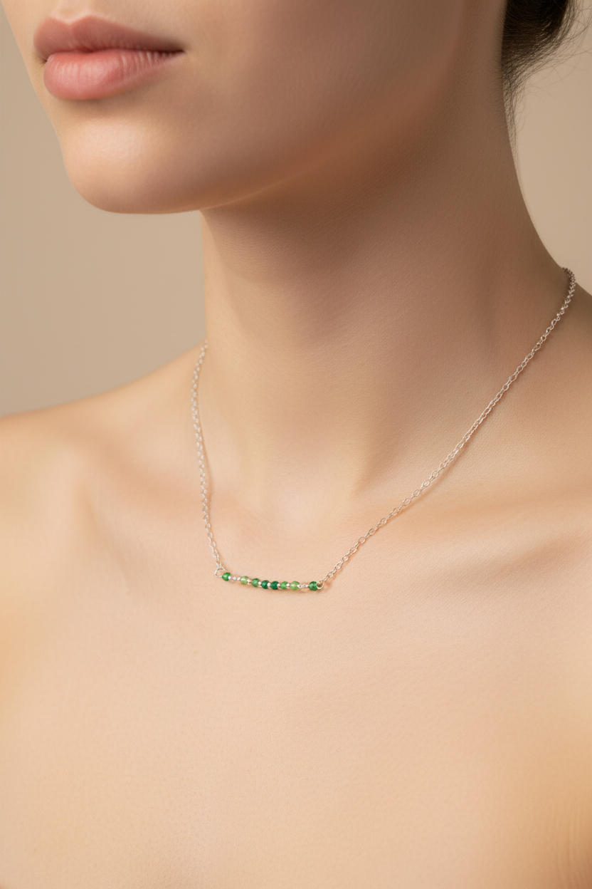 A close-up of a model's neck and upper chest wearing a delicate silver-toned bar necklace with small green gemstones, set against a soft beige background.