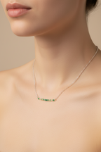 A close-up of a model's neck and upper chest wearing a delicate silver-toned bar necklace with small green gemstones, set against a soft beige background.