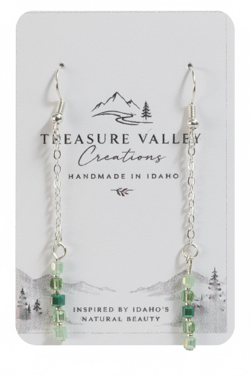 Treasure Valley Creations handmade dangle earrings with silver chains, clear faceted beads, and light green beads, displayed on a white card backing with the brand logo.