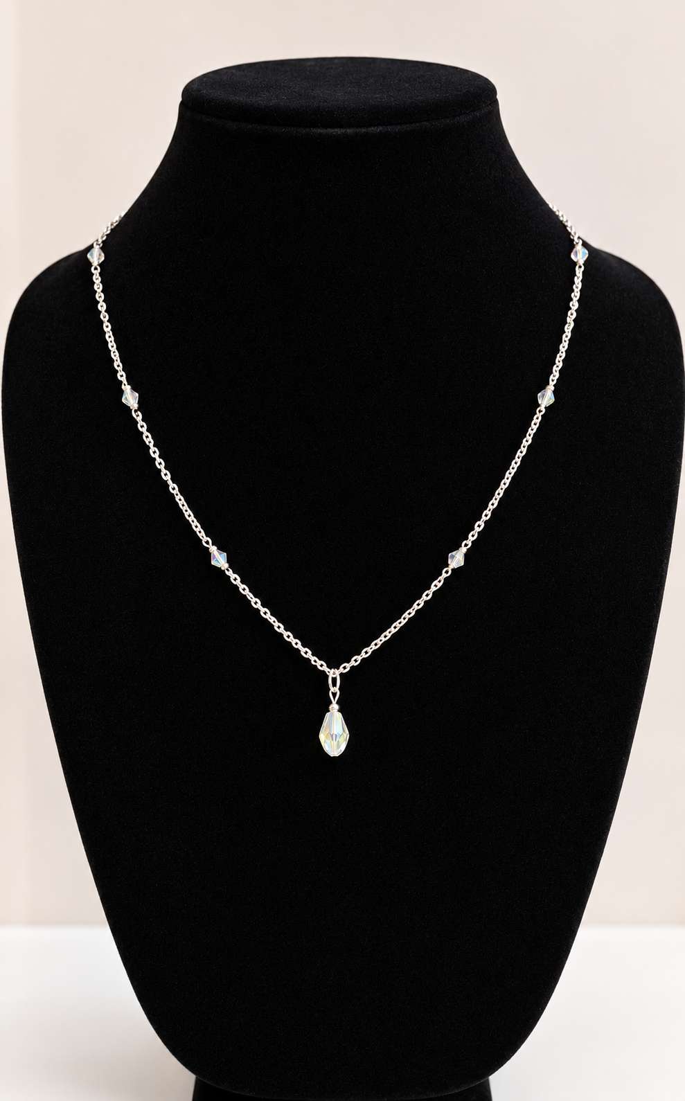 A delicate silver chain necklace with a teardrop-shaped iridescent pendant, displayed on a black velvet jewelry bust against a light beige background.