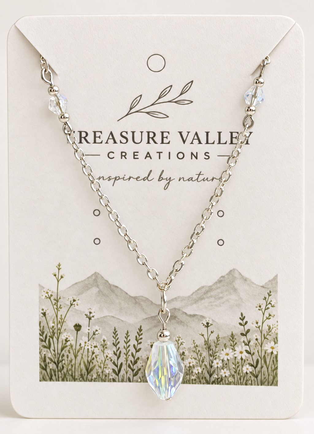 A silver chain necklace with a faceted iridescent crystal pendant, displayed on a cream-colored jewelry card from Treasure Valley Creations.