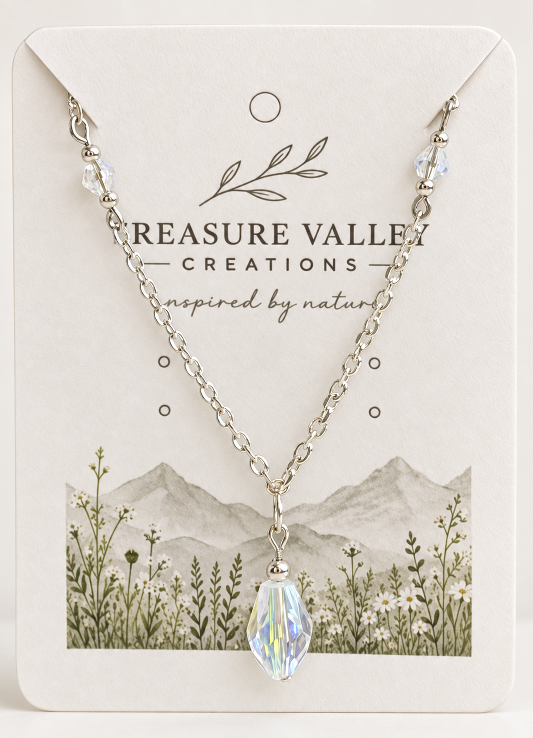 A silver chain necklace with a faceted iridescent crystal pendant, displayed on a cream-colored jewelry card from Treasure Valley Creations.