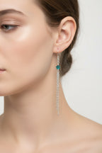A pair of silver dangle earrings with small green and yellow gemstone accents, hanging against a plain light gray background.