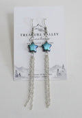 A pair of silver-toned dangle earrings with small green bead accents, displayed on a white branded card against a light gray background.
