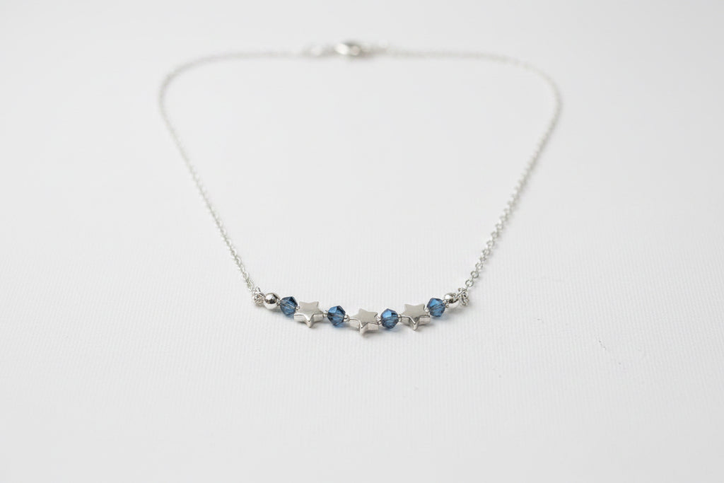 A silver chain necklace featuring a central row of small silver stars and three dark blue faceted gemstones, displayed on a textured off-white fabric background.