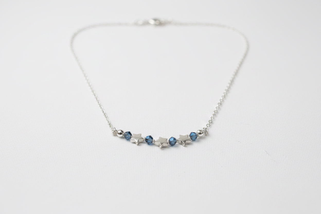 A silver chain necklace featuring a central row of small silver stars and three dark blue faceted gemstones, displayed on a textured off-white fabric background.
