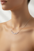 A close-up of a model wearing a delicate silver necklace with a curved row of clear, sparkling gemstones at the center, set against a soft, light-toned background.