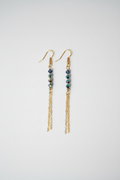 A pair of gold dangle earrings with small dark blue beads and iridescent green accents, hanging against a plain light gray background.