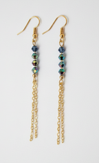 A pair of gold dangle earrings with small dark blue beads and iridescent green accents, hanging against a plain light gray background.