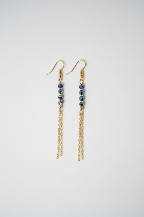 A pair of gold dangle earrings with small dark blue beads and iridescent green accents, hanging against a plain light gray background.