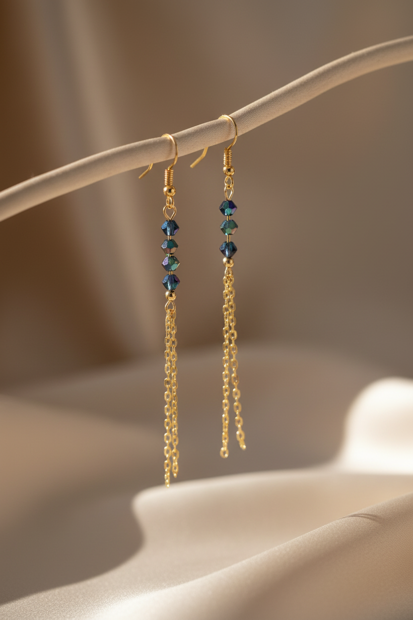 A pair of gold dangle earrings with small iridescent blue beads and long gold chain drops, hanging from a curved cream display stand against a softly lit beige background.