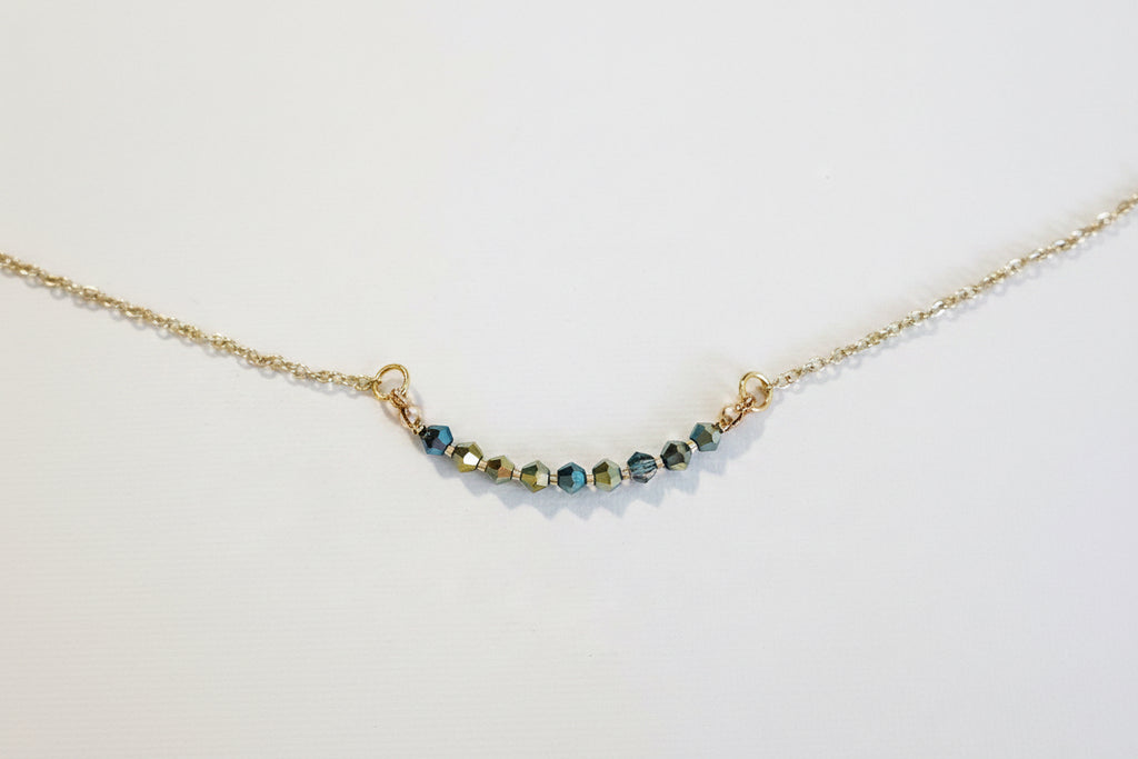 A delicate gold chain necklace featuring a short, curved bar of faceted beads in blue, green, and gold, displayed on a clean white background.