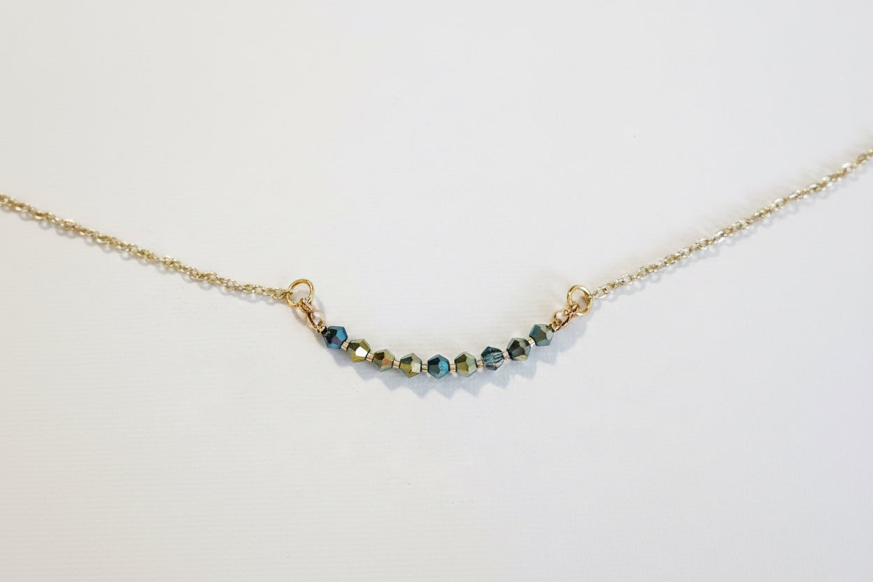 A delicate gold chain necklace featuring a short, curved bar of faceted beads in blue, green, and gold, displayed on a clean white background.