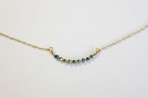 A delicate gold chain necklace featuring a short, curved bar of faceted beads in blue, green, and gold, displayed on a clean white background.