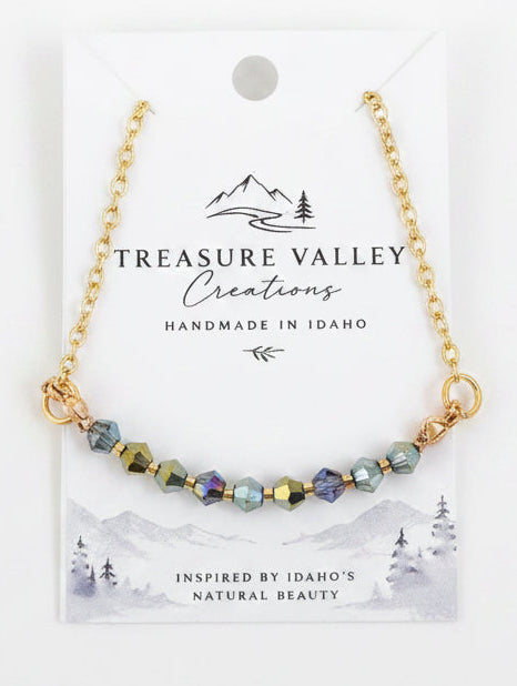 A delicate gold chain necklace with a row of small, iridescent green and gold beads, displayed on a white backing card from Treasure Valley Creations.
