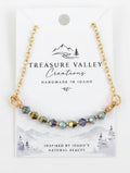 A delicate gold chain necklace with a row of small, iridescent green and gold beads, displayed on a white backing card from Treasure Valley Creations.