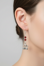 A close-up of a model's ear wearing a dangling earring with silver-toned metal, small red beads, and a swan-shaped charm.