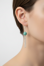 A close-up of a model's ear wearing a dangling earring with a round turquoise-green bead and a silver-toned tassel charm.
