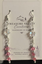 A pair of dangling earrings displayed on a branded card. The earrings have silver-toned hooks and beads, clear faceted star-shaped beads, and pink faceted round beads at the bottom.