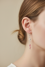 A close-up side profile of a woman wearing a long silver dangle earring with pink faceted beads and clear crystal details, against a soft beige background.