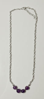 A silver chain necklace with three purple beads at the center, laid flat on a light gray surface.