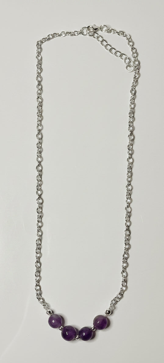 A silver chain necklace with three purple beads at the center, laid flat on a light gray surface.