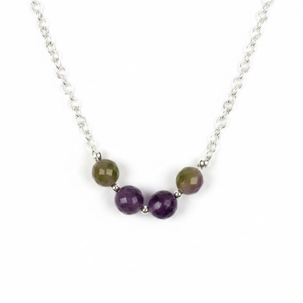 A close-up of a silver chain necklace featuring five round beads: three faceted purple beads and two larger oval beads with a brown and green marbled pattern, separated by small silver spacer beads, on a plain light gray background.