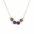 A close-up of a silver chain necklace featuring five round beads: three faceted purple beads and two larger oval beads with a brown and green marbled pattern, separated by small silver spacer beads, on a plain light gray background.