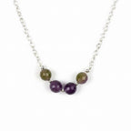 A close-up of a silver chain necklace featuring five round beads: three faceted purple beads and two larger oval beads with a brown and green marbled pattern, separated by small silver spacer beads, on a plain light gray background.