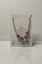 A silver chain necklace with four purple and olive-green gemstone beads, displayed on its white backing card which shows the brand 'Treasure Valley Creations'.