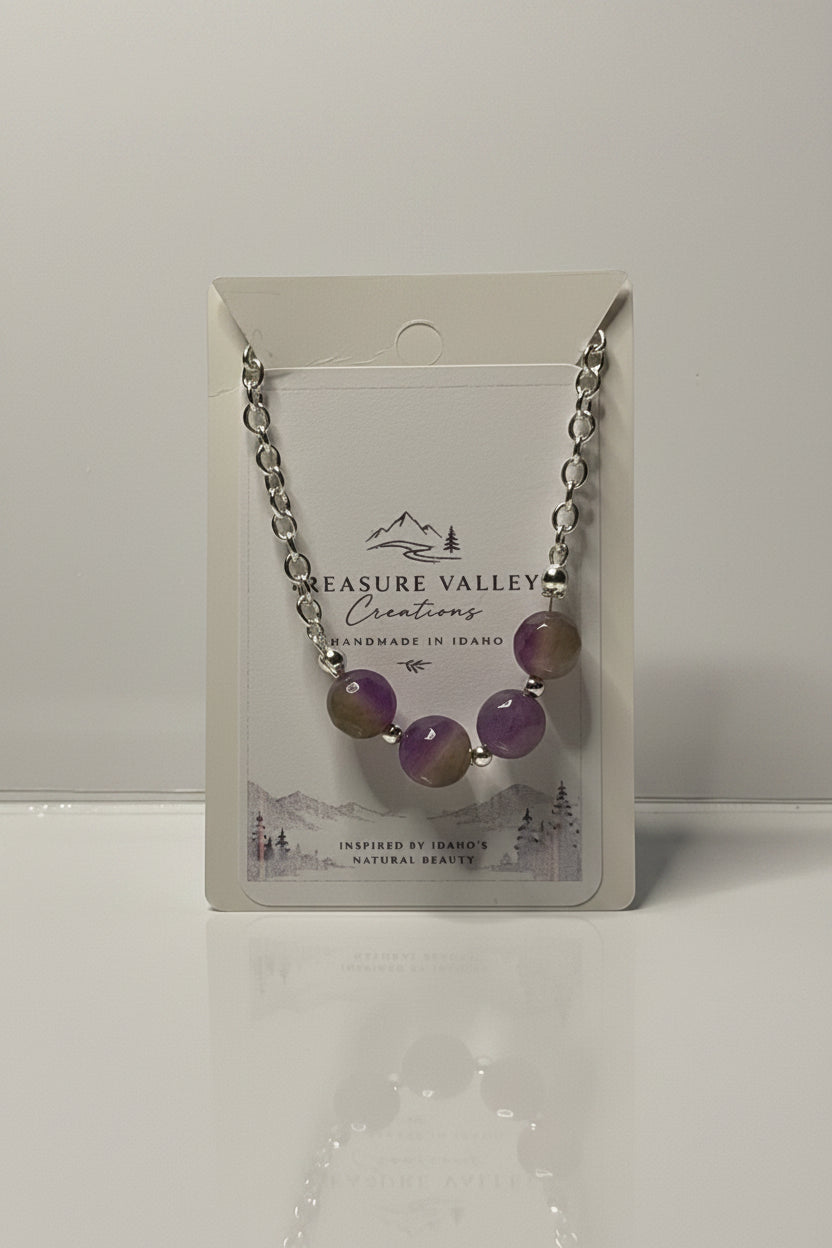 A silver chain necklace with four purple and olive-green gemstone beads, displayed on its white backing card which shows the brand 'Treasure Valley Creations'.
