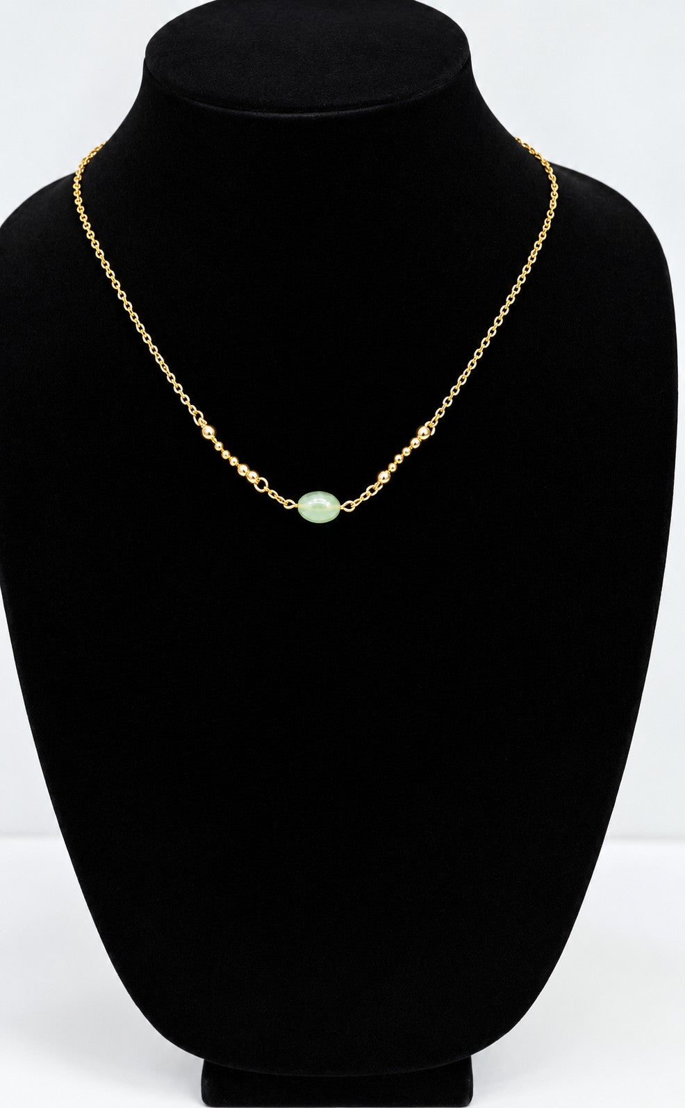 A delicate gold chain necklace with a single light green oval stone pendant, displayed on a black velvet jewelry bust against a white background.