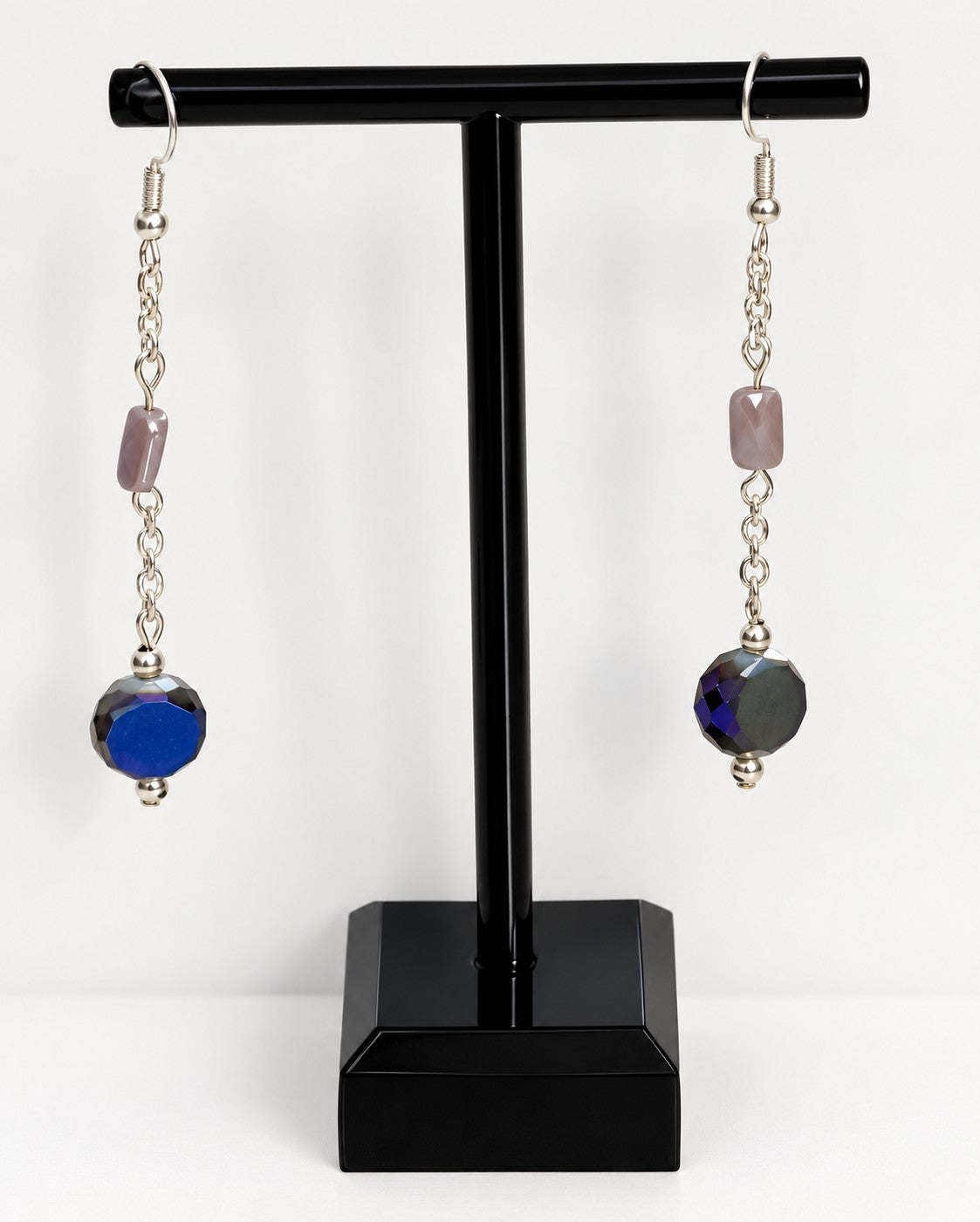 A pair of long dangling earrings displayed on a black stand against a white background. Each earring has silver-tone chain links, a small dusty pink rectangular bead, and a larger faceted blue bead at the bottom.