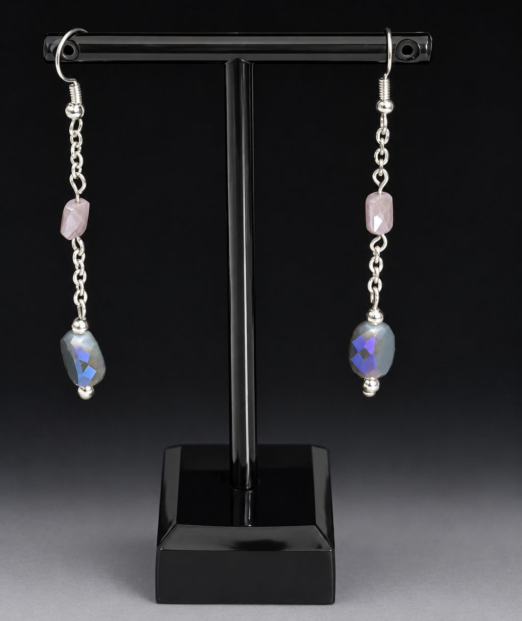 A pair of silver dangle earrings displayed on a black T-stand against a dark studio background. Each earring has a small pink stone, a silver chain, and a larger faceted blue-purple stone with a triangular cutout.