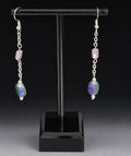 A pair of silver dangle earrings displayed on a black T-stand against a dark studio background. Each earring has a small pink stone, a silver chain, and a larger faceted blue-purple stone with a triangular cutout.